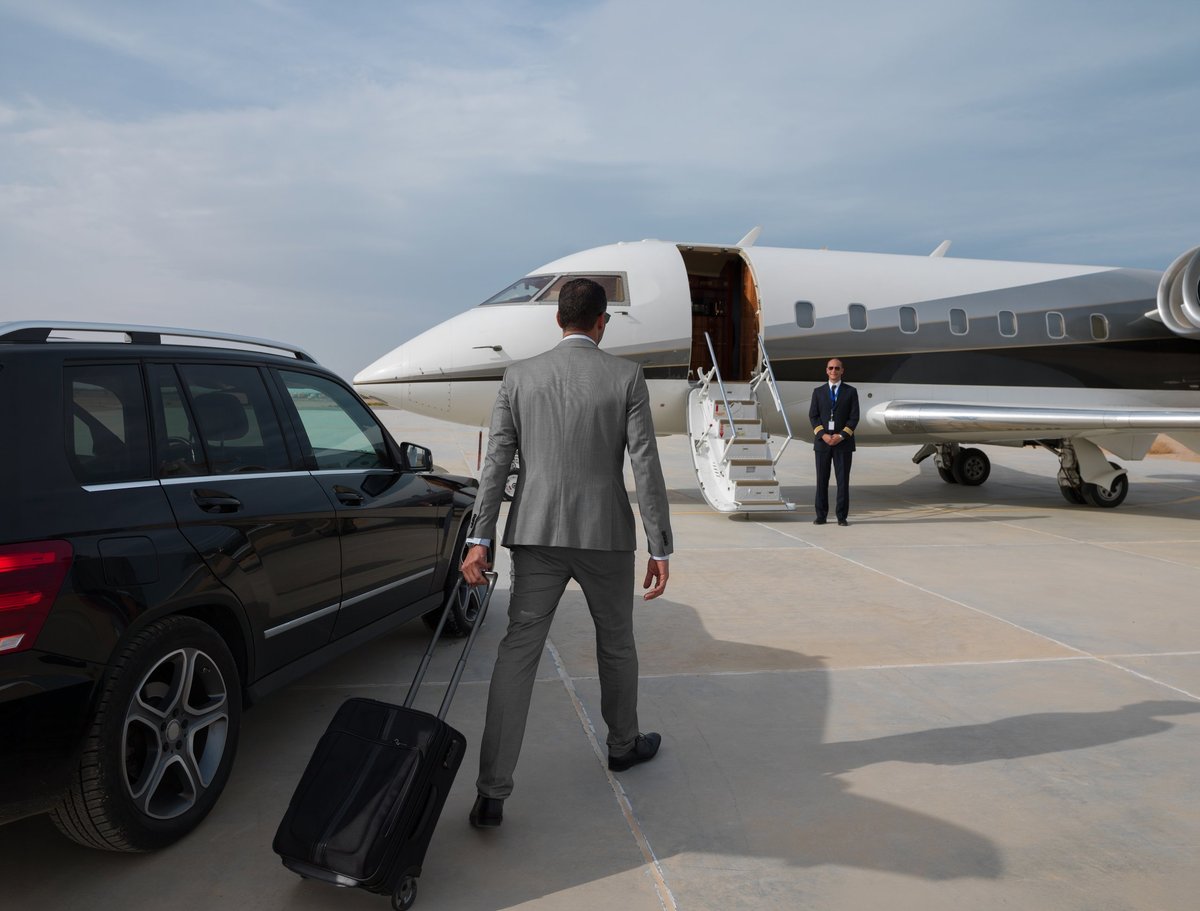 Business executives walking across a sunlit tarmac to board a private jet, with a luxury SUV and attendant waiting